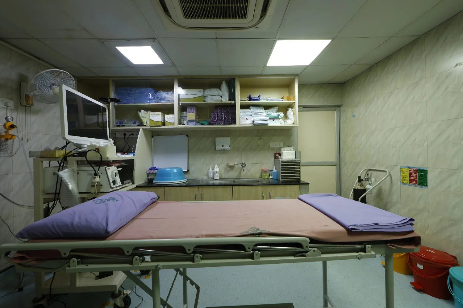 An empty patient bed in an endoscopy procedure room with medical equipment and shelving.