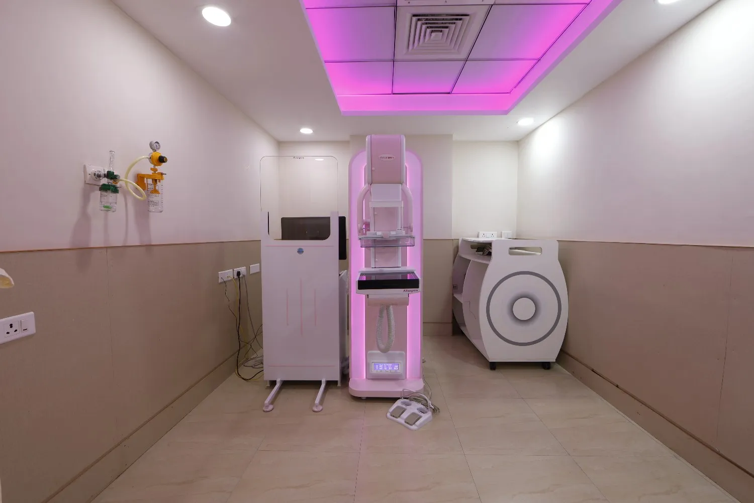 An empty mammogram room featuring a pink-lit mammography machine and protective shield.