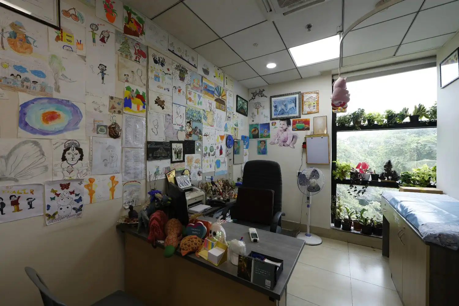 An empty pediatric doctor's room with children's drawings on the wall, a desk with toys, and an examination bed.