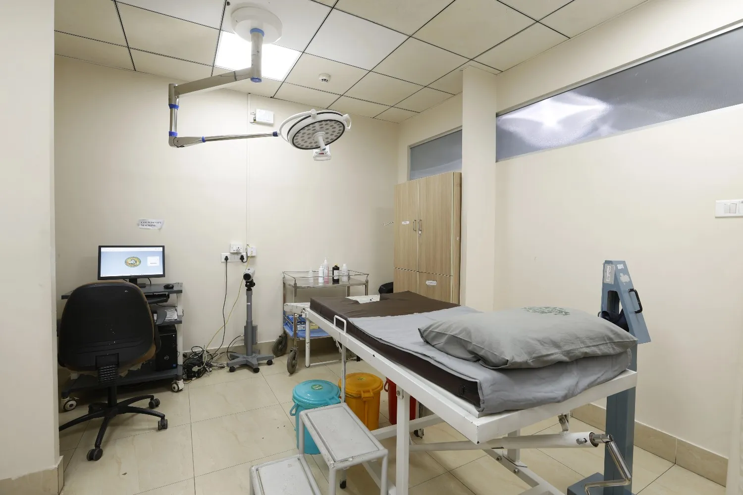 An empty gynecologist examination room with an examination table, overhead light, and a computer desk.