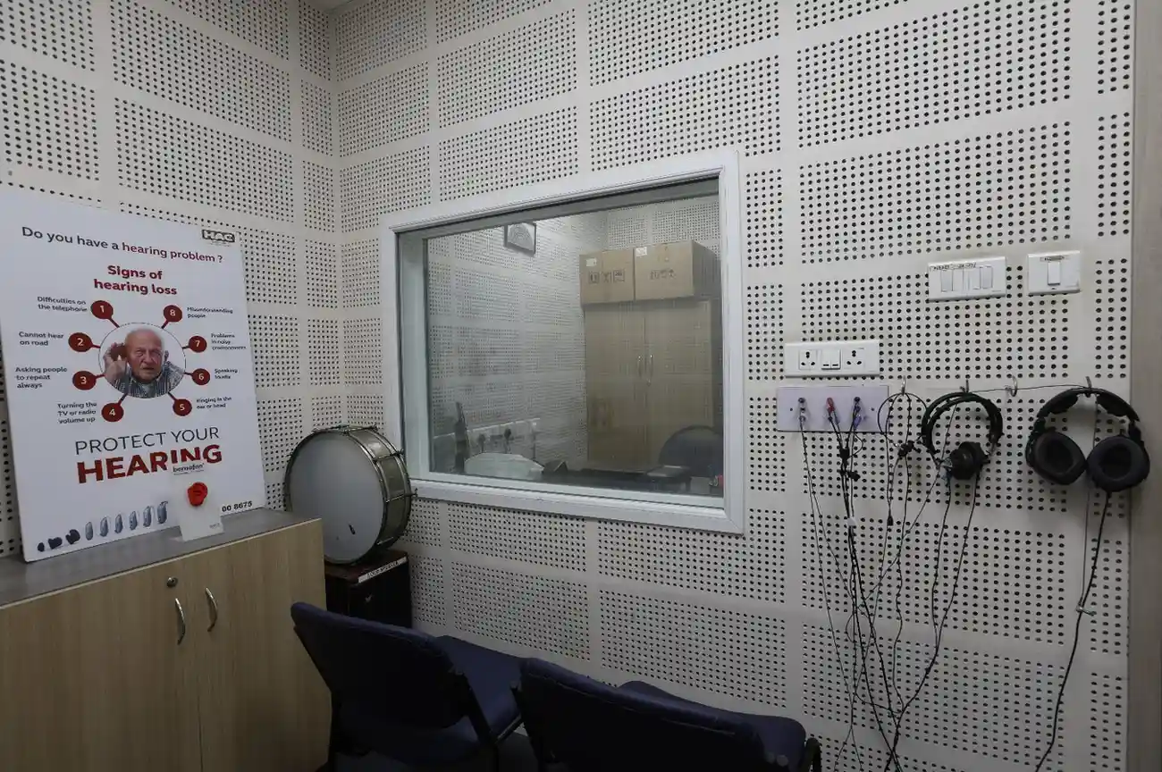 An empty audiology room with acoustic wall panels, a window to a testing booth, and hanging headphones.